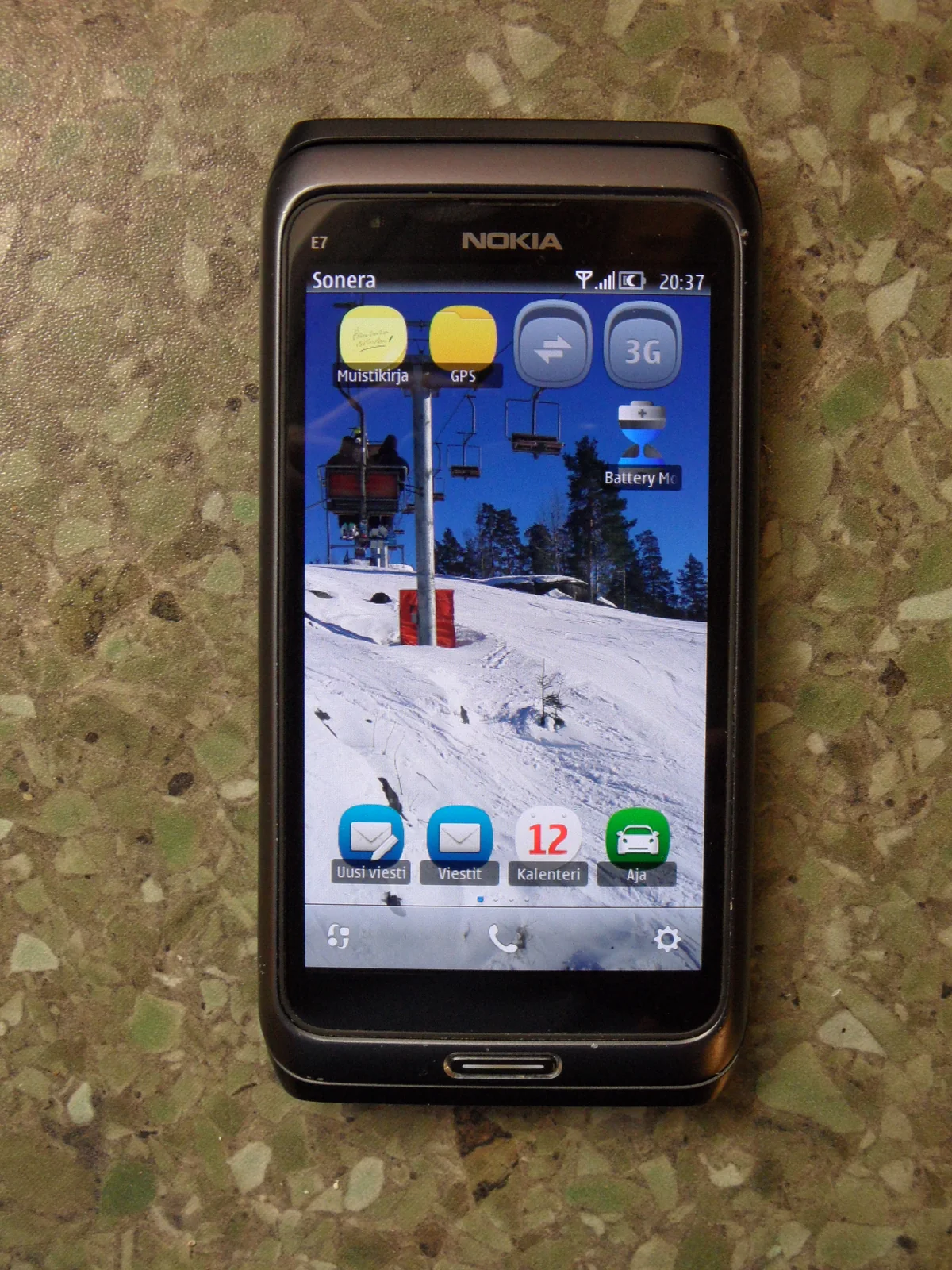 A Nokia E7-00 with its slider closed, showing the Symbian home screen